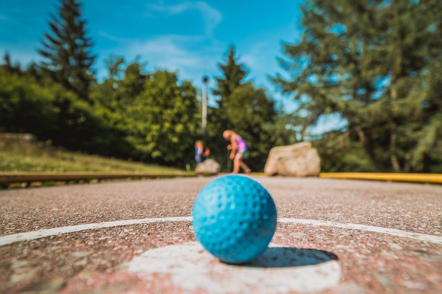 im Vordergrund Minigolf auf einer Minigolfbahn, im Hintergrund eine Person, Bäume, blauer Himmel